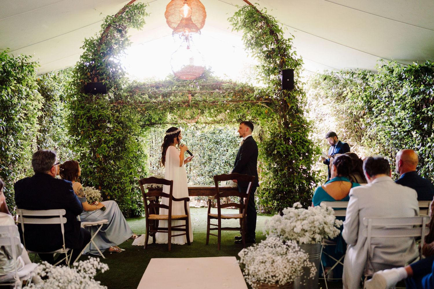 Wide shot of bride and groom exchanging vows under a green ivy arch at outdoor wedding.