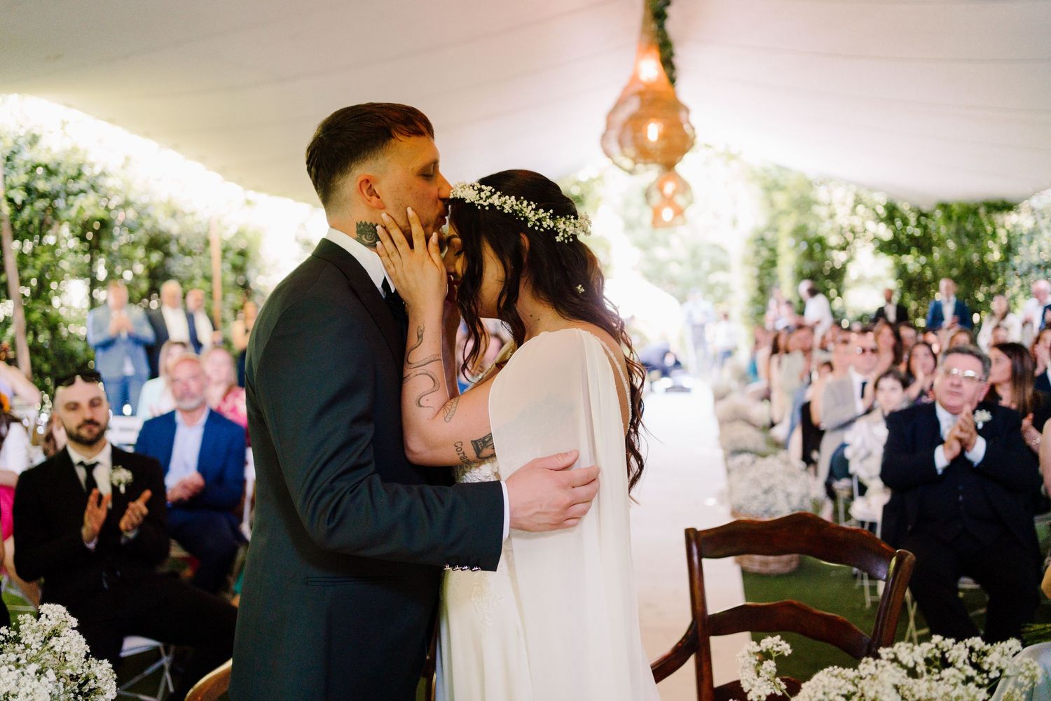 Bride and groom share their first kiss as newlyweds while guests applaud at wedding reception.