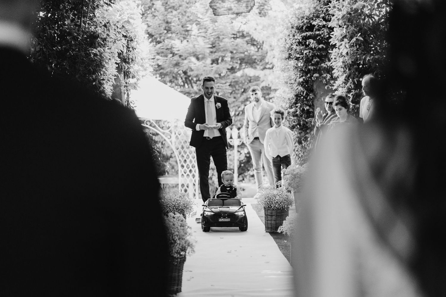 Black and white photo of groom arriving at outdoor wedding ceremony on a riding lawn mower.