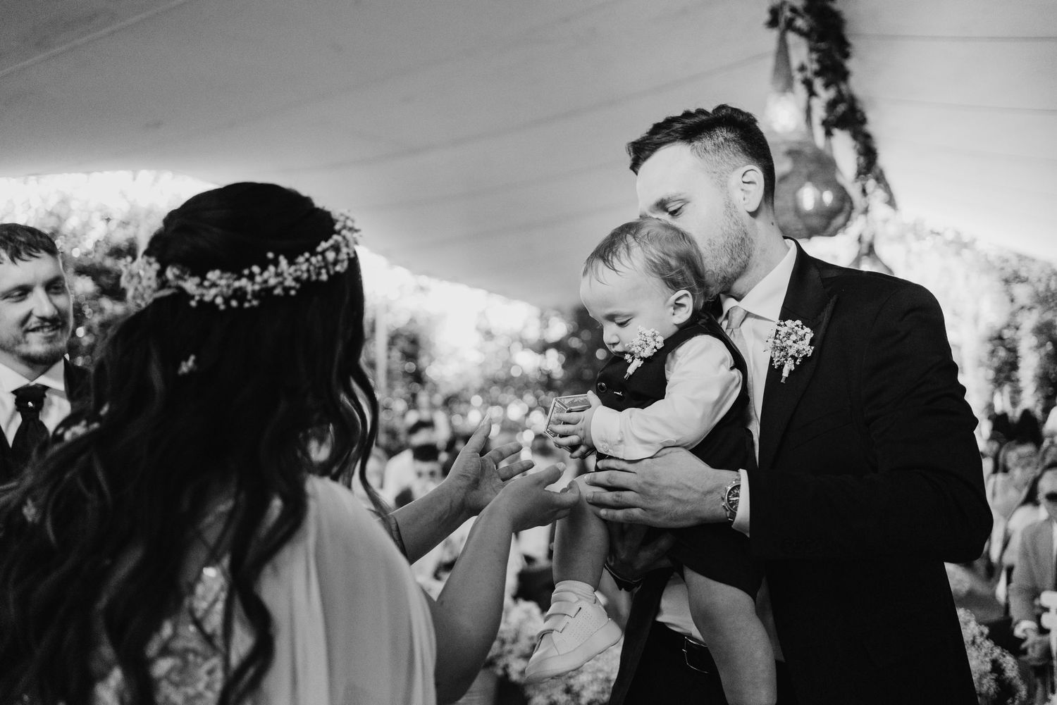 Groom holding baby while bride places ring during outdoor wedding ceremony under white tent.