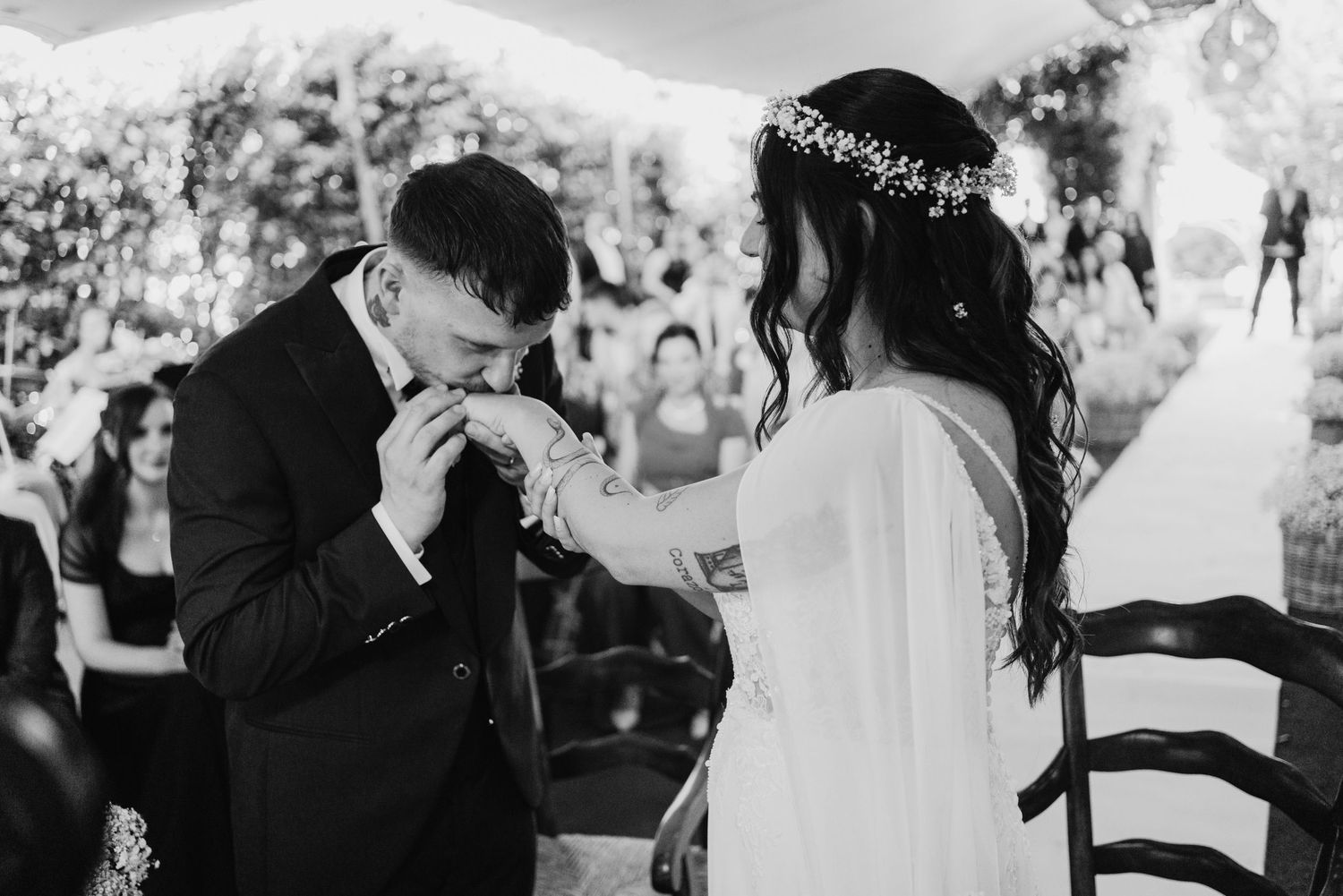 Groom kisses bride's hand tenderly during black and white wedding ceremony moment.