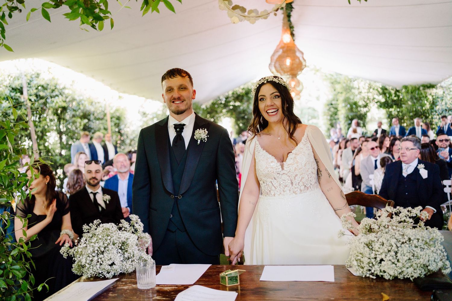 Bride and groom smile while signing wedding register at floral-decorated outdoor ceremony.