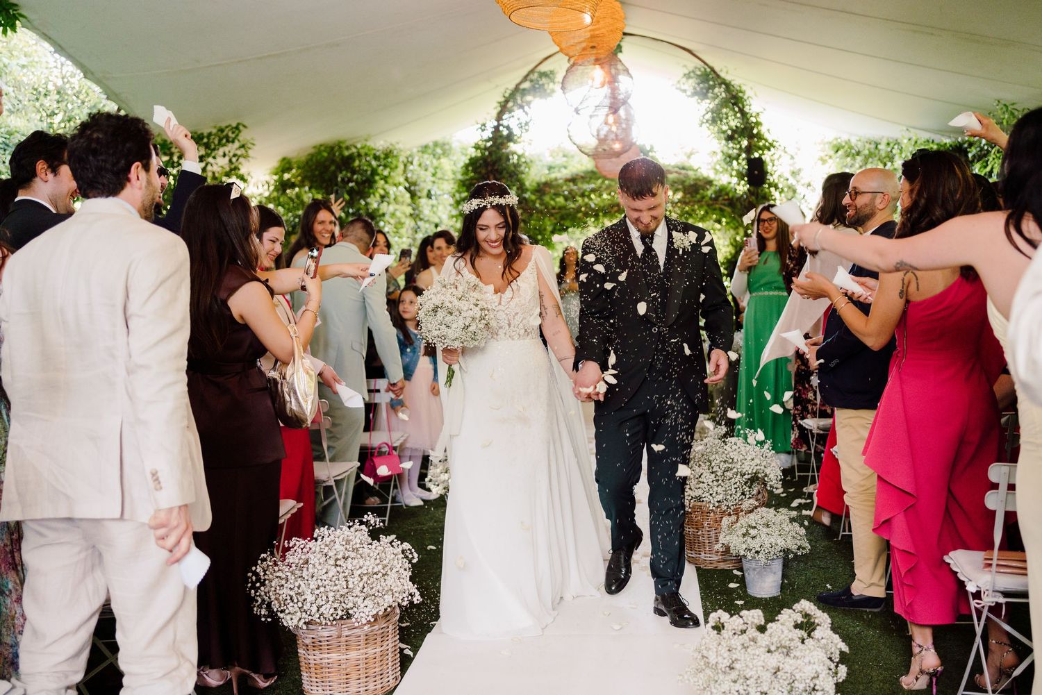 Bride and groom exit ceremony as colorful guests celebrate with confetti in garden venue.