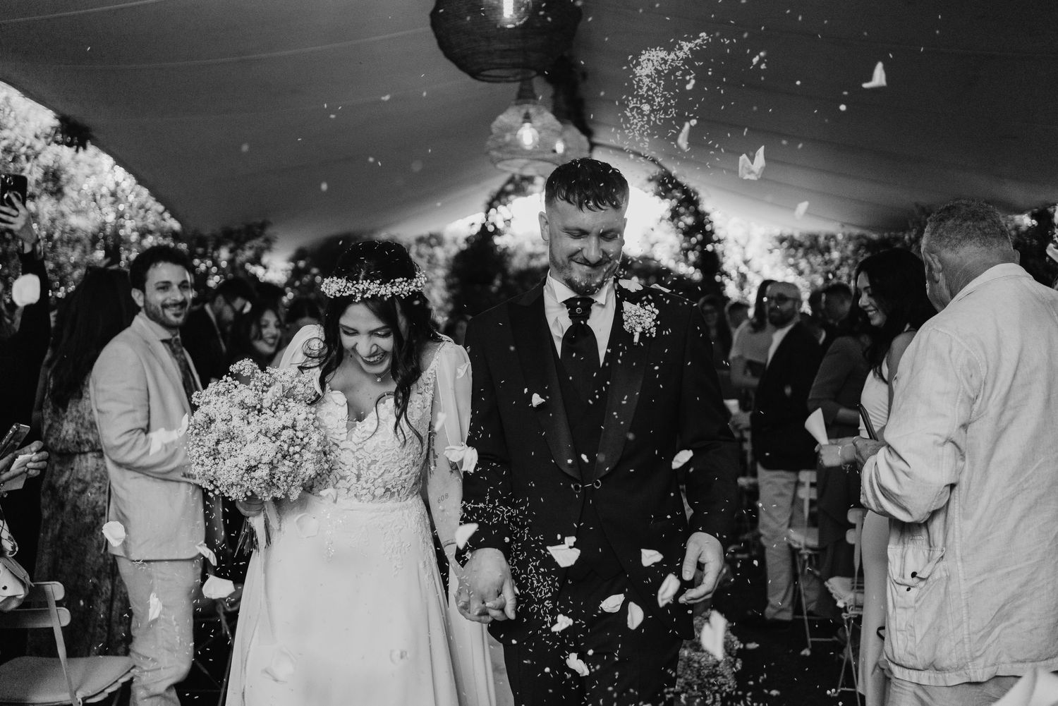 Bride and groom walk through confetti shower in black and white outdoor wedding photo.