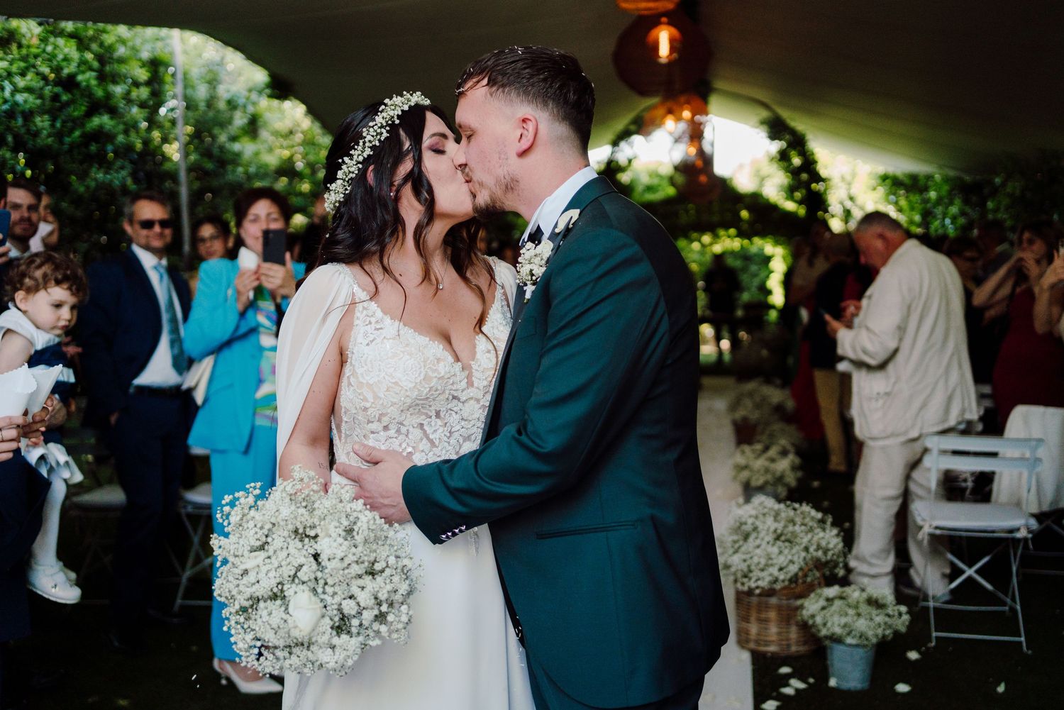 Bride and groom share romantic first kiss holding bouquet at outdoor garden wedding.