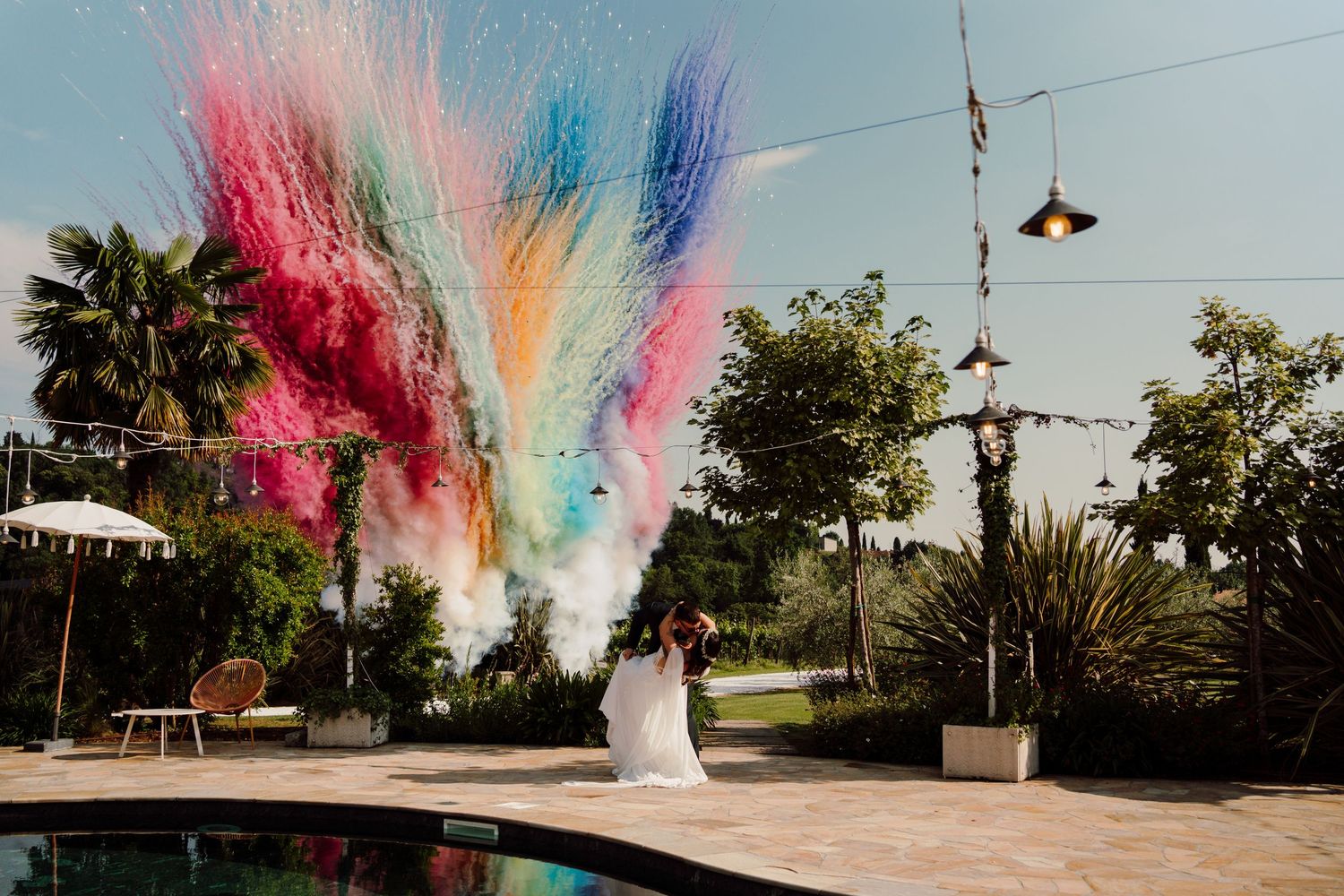 Bride and groom celebrate with vibrant colorful powder explosion near outdoor pool.