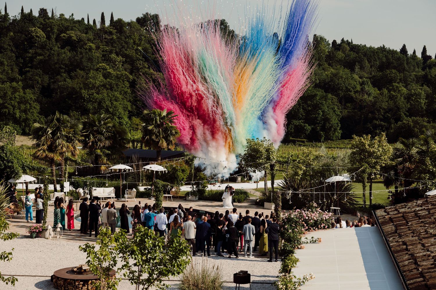 Colorful powder explosion fills the sky above wedding guests at an outdoor ceremony in a lush garden venue.