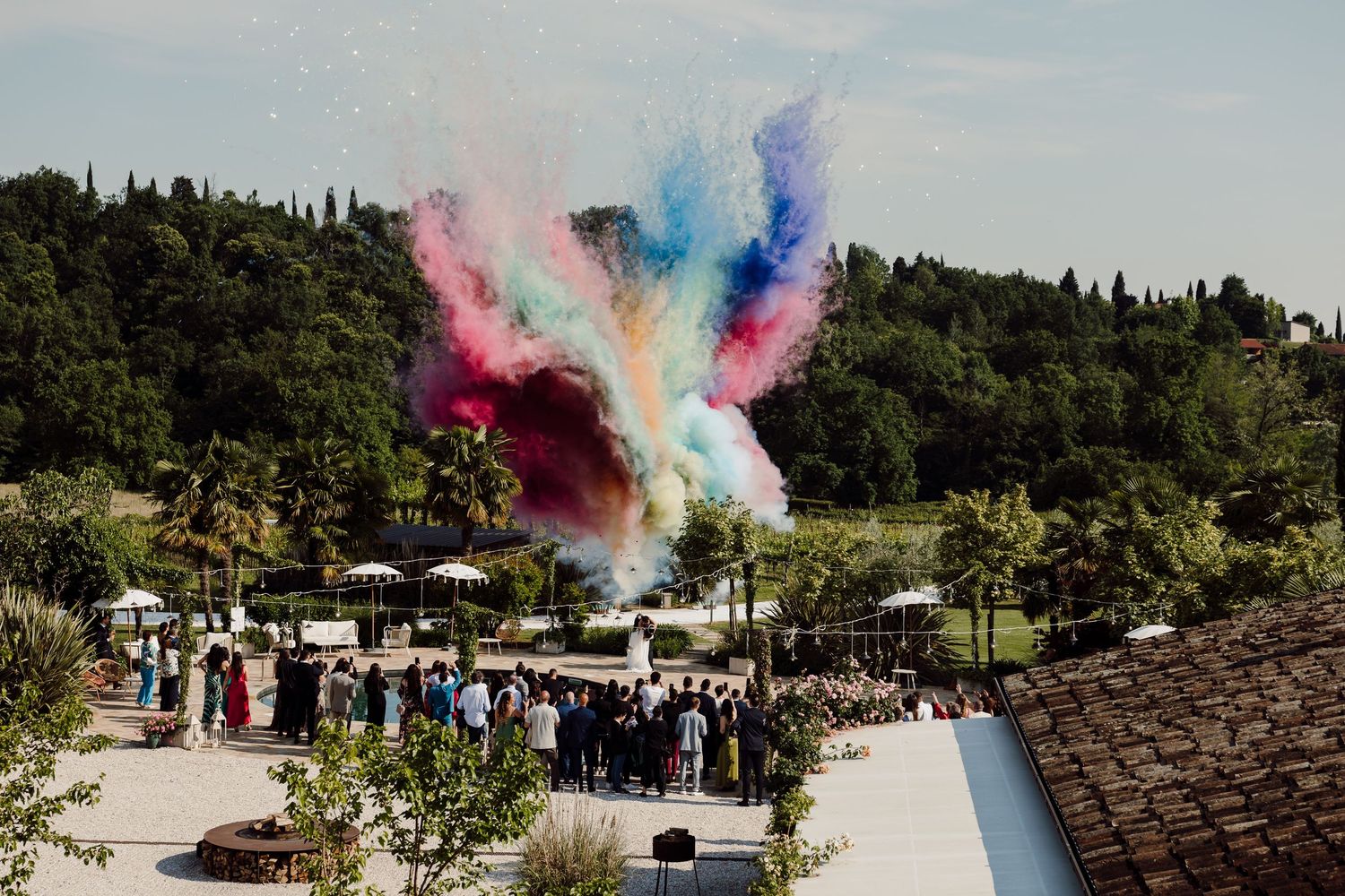 Vibrant multicolored powder burst above a crowd of wedding guests celebrating outdoors in a scenic garden.