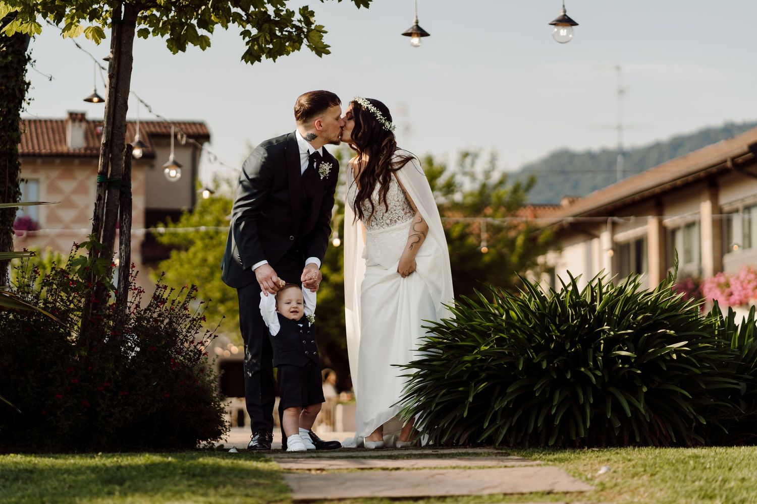 Bride and groom share a kiss while holding a young child between them in a sunny garden setting.