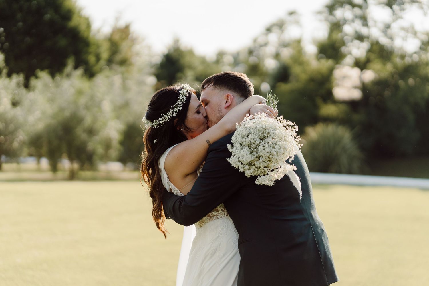 Bride and groom share a romantic embrace and kiss outdoors on a sunny green lawn after their wedding.