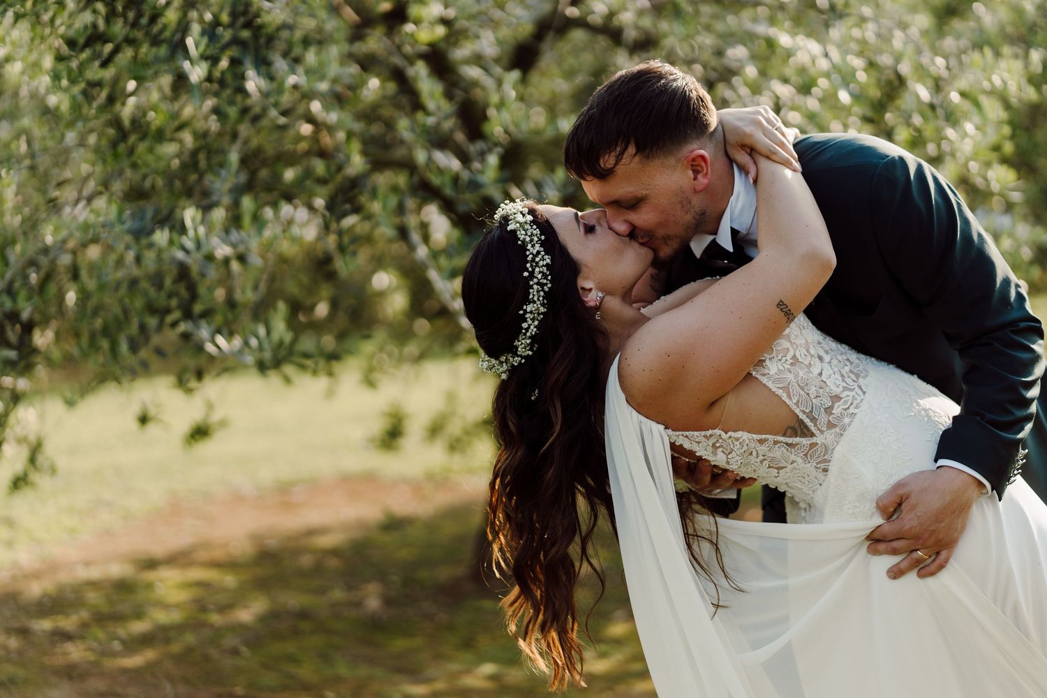 Groom dips bride for a romantic kiss beneath olive trees in a sunlit outdoor wedding portrait.