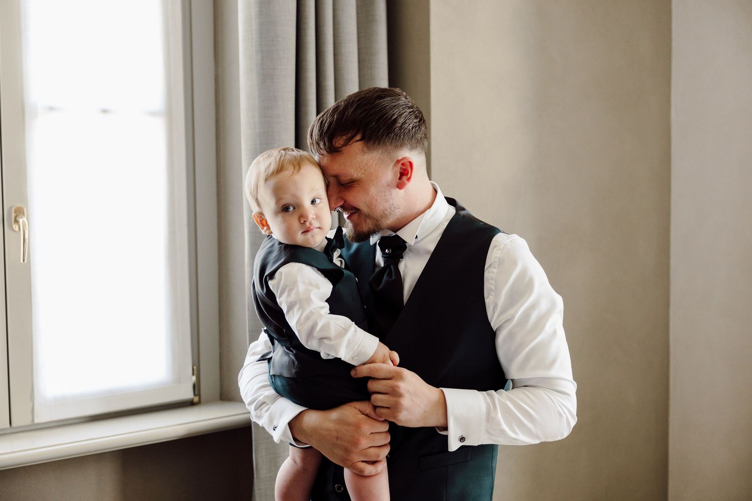 Groom in vest and tie tenderly kissing his baby son by a bright window.