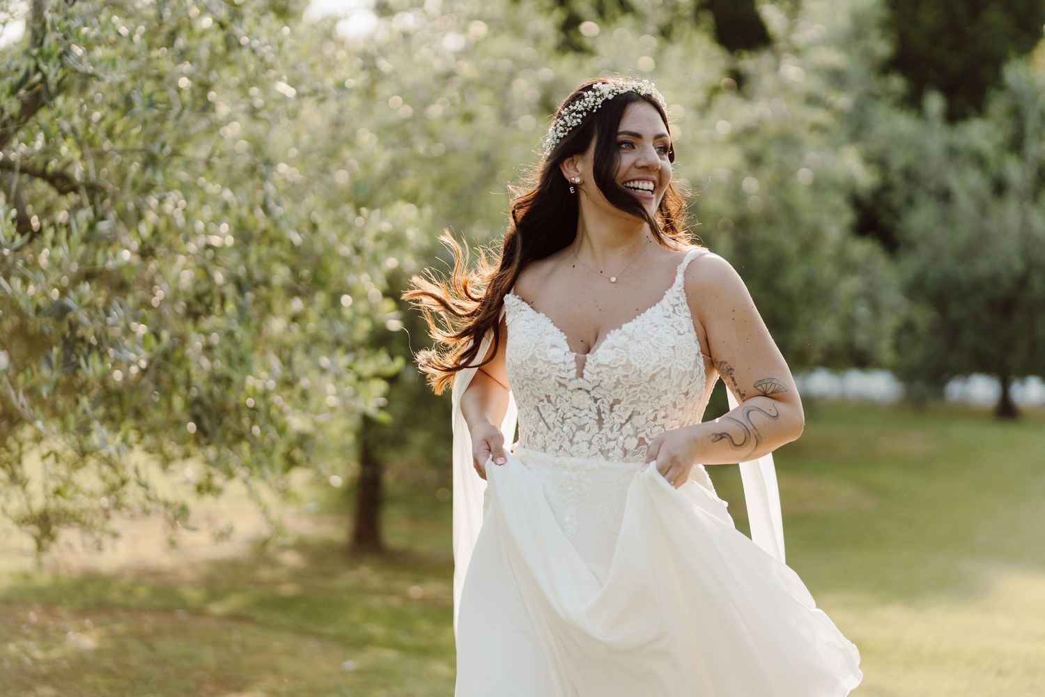 Radiant bride in a lace wedding gown smiles joyfully while walking through a sunlit olive tree garden.