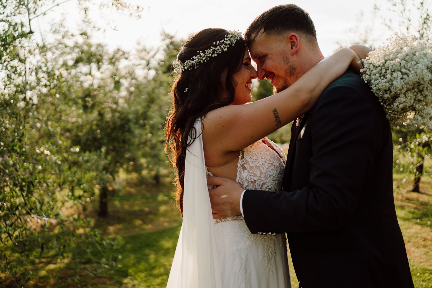 Romantic bride and groom embracing forehead to forehead in a sunlit orchard setting.