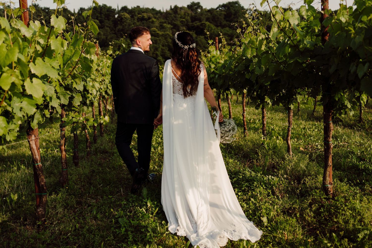 Bride and groom walking hand in hand through lush vineyard rows on their wedding day.