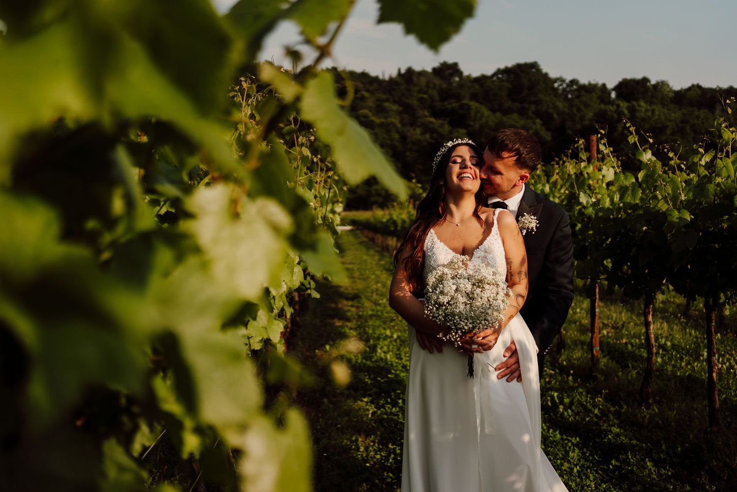Happy bride holding baby's breath bouquet with groom kissing her cheek in vineyard.