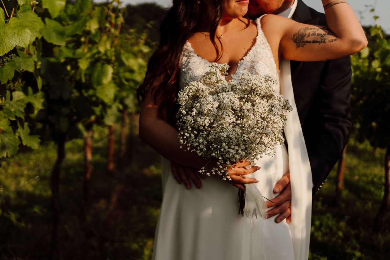 Close-up of bride holding white baby's breath bouquet while groom embraces her in vineyard.