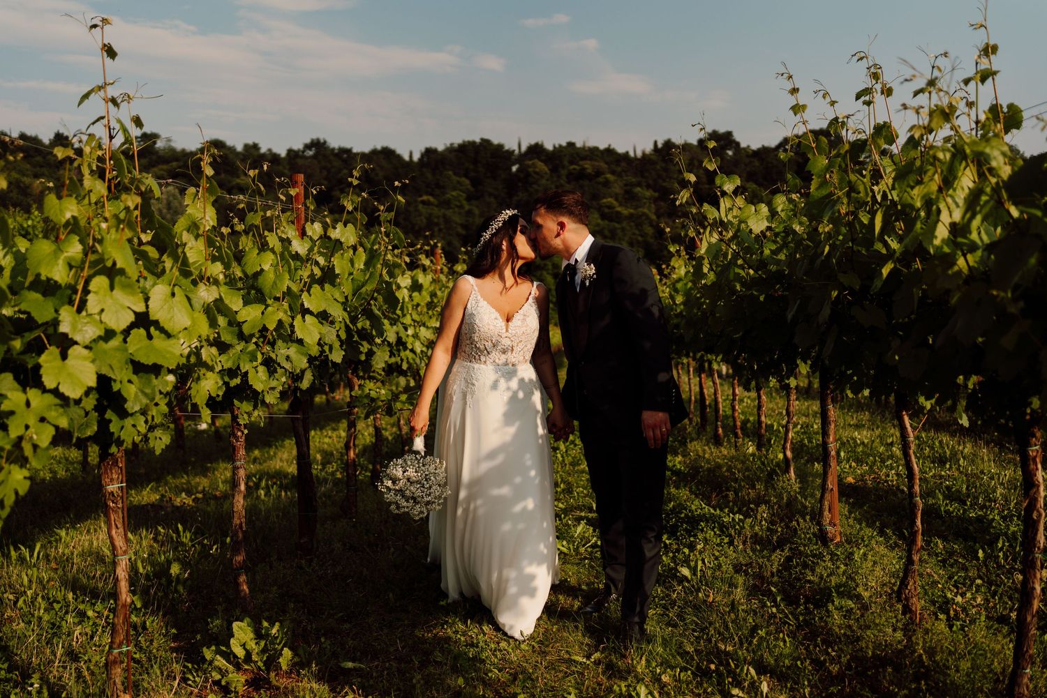 Bride and groom sharing a kiss while strolling through a lush green vineyard at dusk.