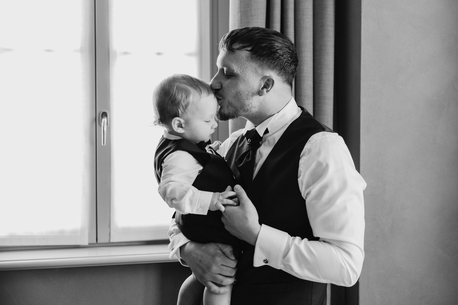 Black and white photo of a dapper groom kissing his smiling baby by a window.