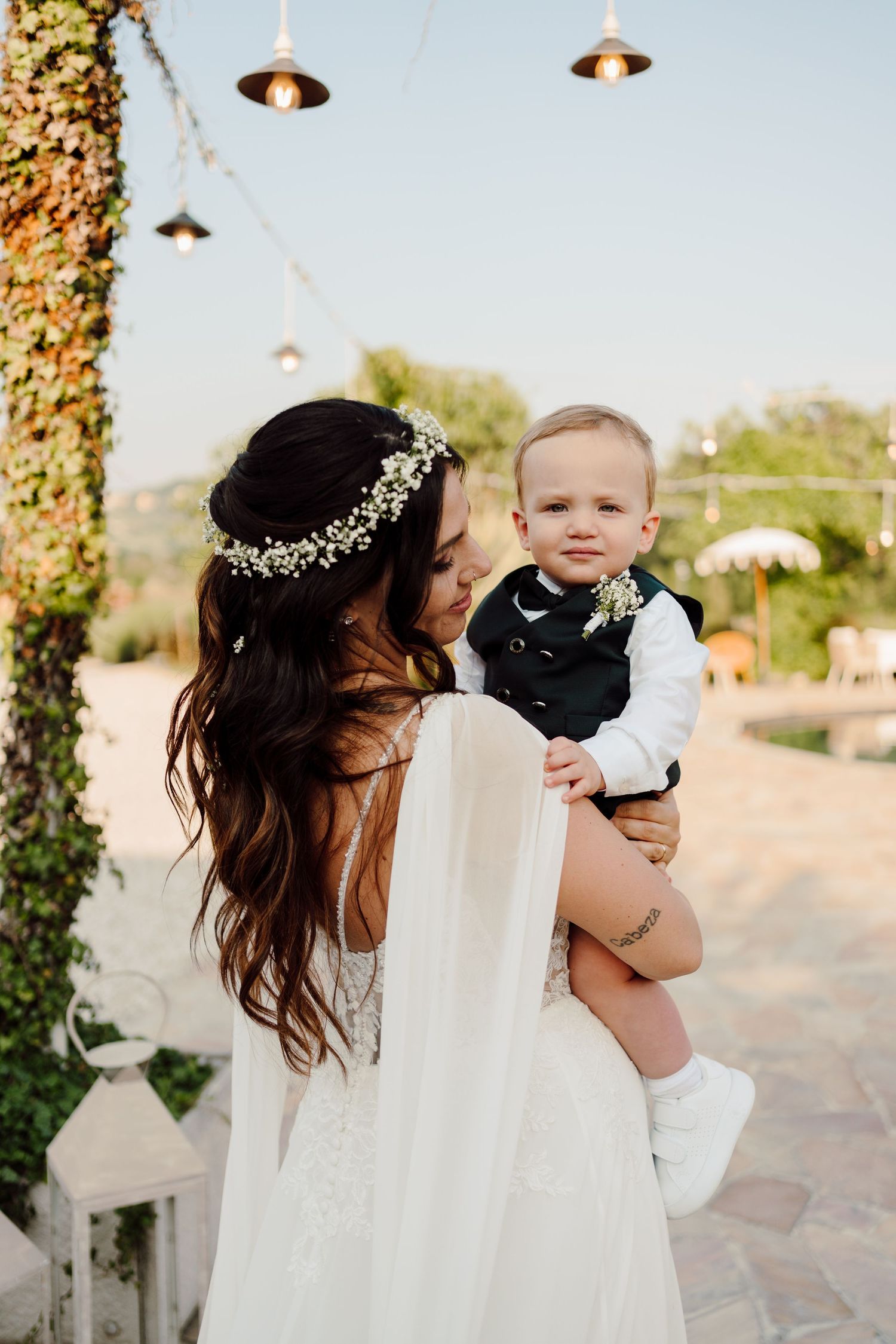 Bride in white gown and floral crown holds smiling baby boy in suit at outdoor wedding venue.