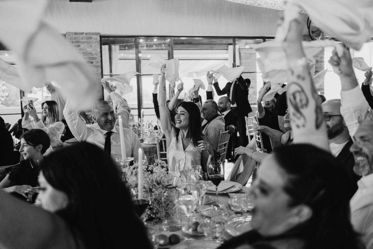 Wedding guests joyfully wave white napkins at elegant reception dinner in black and white photo.