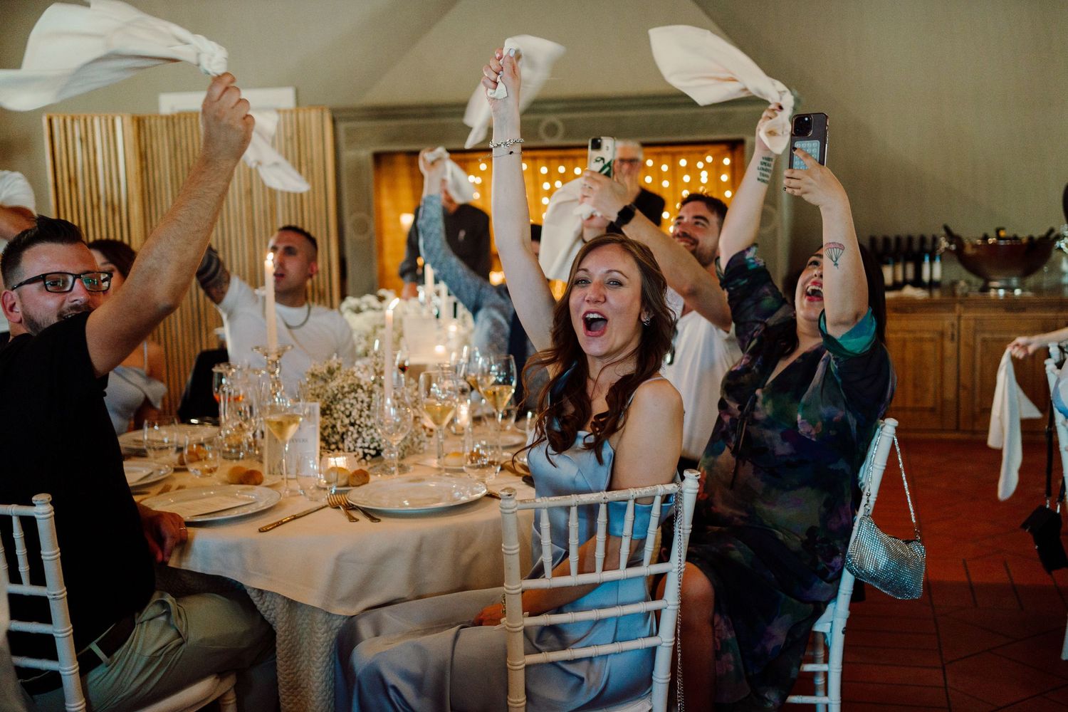 Excited wedding guests wave napkins in celebration at beautifully decorated indoor reception table.