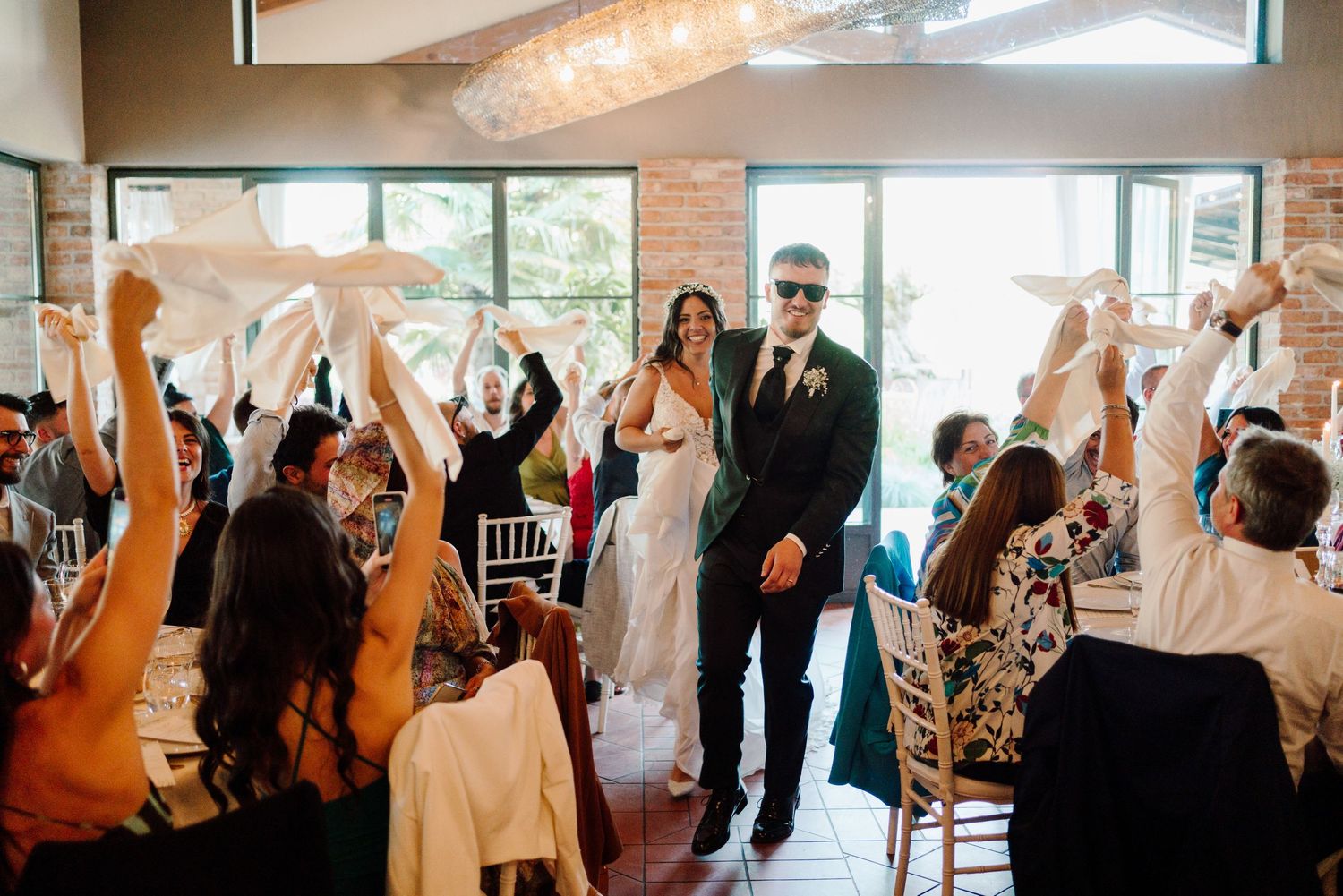 Bride and groom in sunglasses make grand entrance as guests wave napkins at wedding reception.
