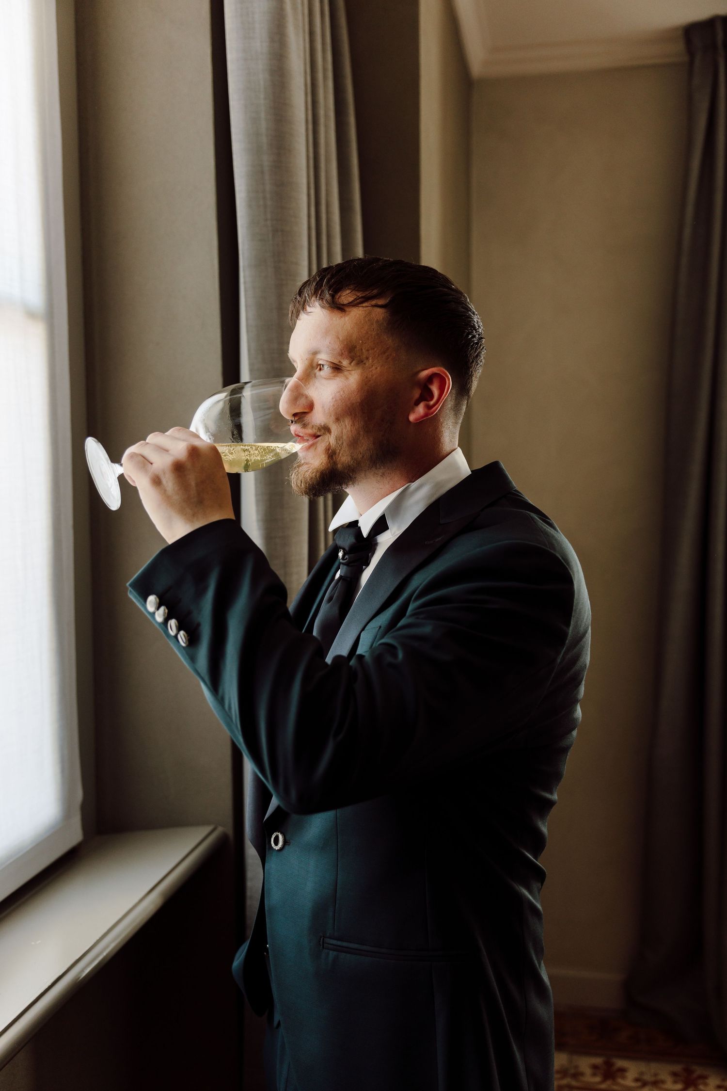 Handsome groom in a dark suit sipping champagne from a glass by a window.
