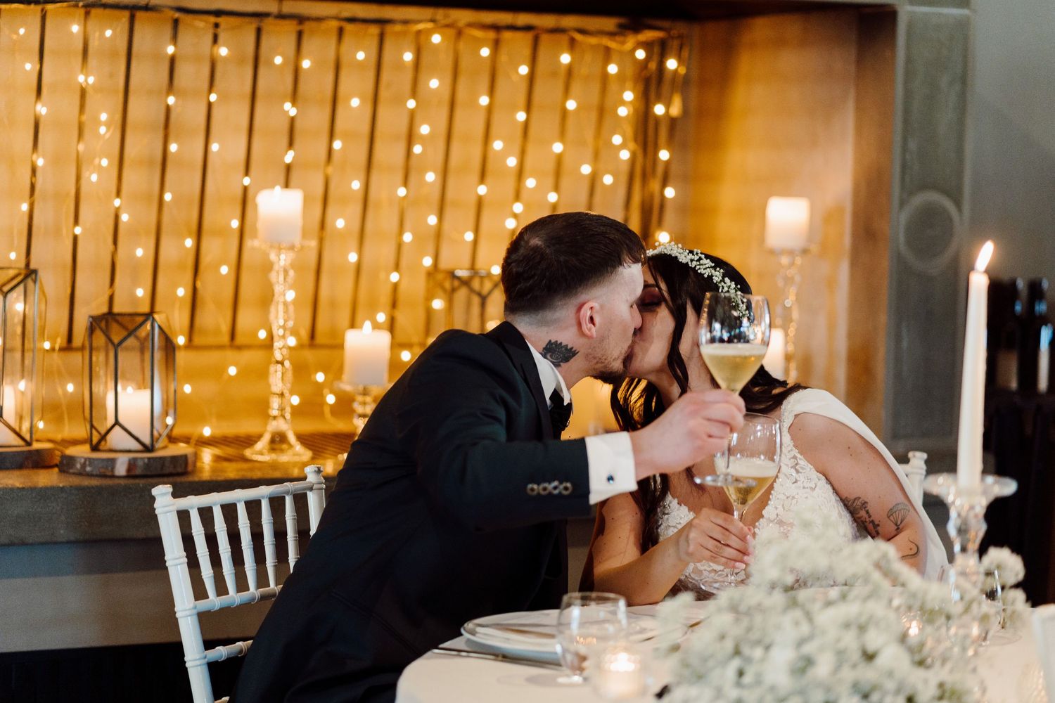 Bride and groom share romantic kiss while toasting champagne at candlelit sweetheart table.