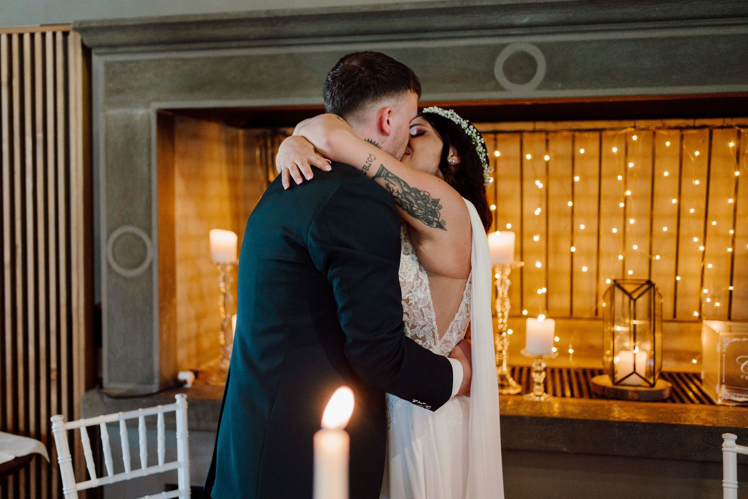Bride and groom share first kiss in front of warm fairy light backdrop at wedding reception.