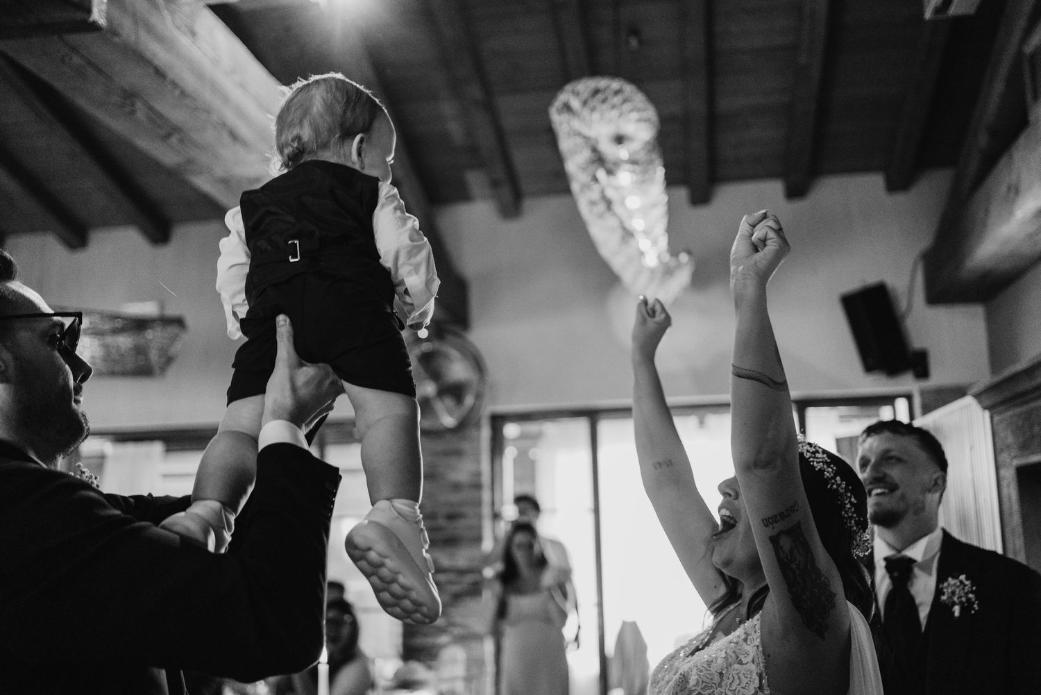 Wedding guest lifts baby in suit while bride celebrates joyfully on dance floor.