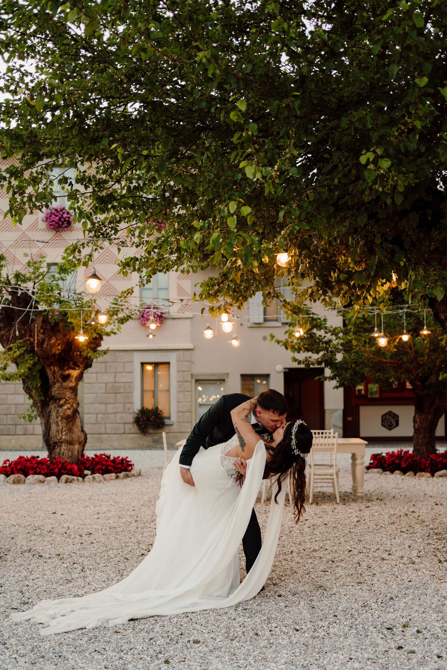 Bride and groom share romantic dip kiss under glowing string lights in garden at dusk.