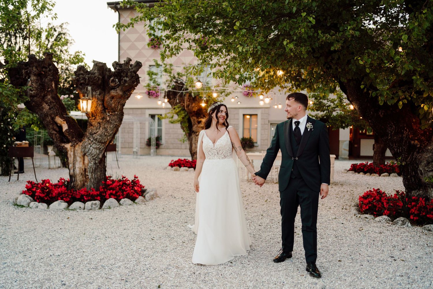Bride and groom hold hands walking through flower-lined gravel courtyard at dusk wedding venue.