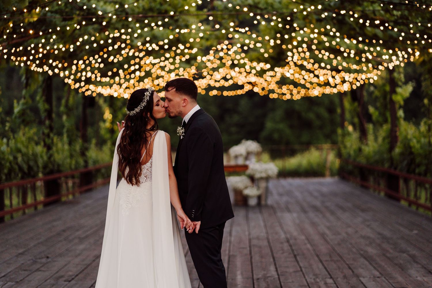 Bride and groom share a romantic moment under a canopy of warm fairy lights on a wooden walkway.