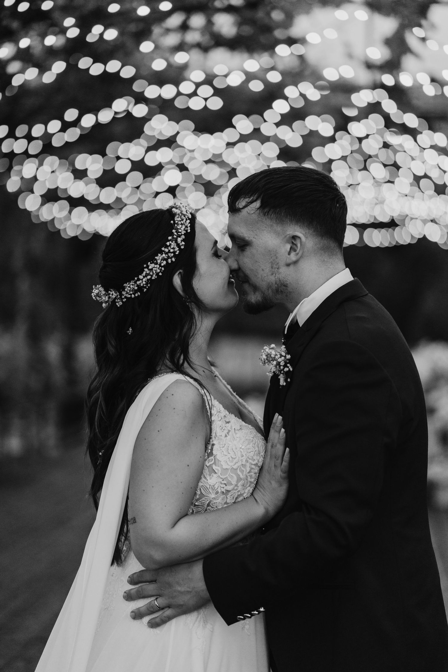 Black and white photo of bride and groom sharing a kiss with bokeh fairy lights glowing behind them.