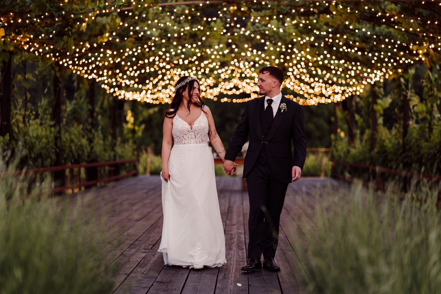 Bride and groom walk hand in hand under a stunning canopy of twinkling fairy lights at dusk.