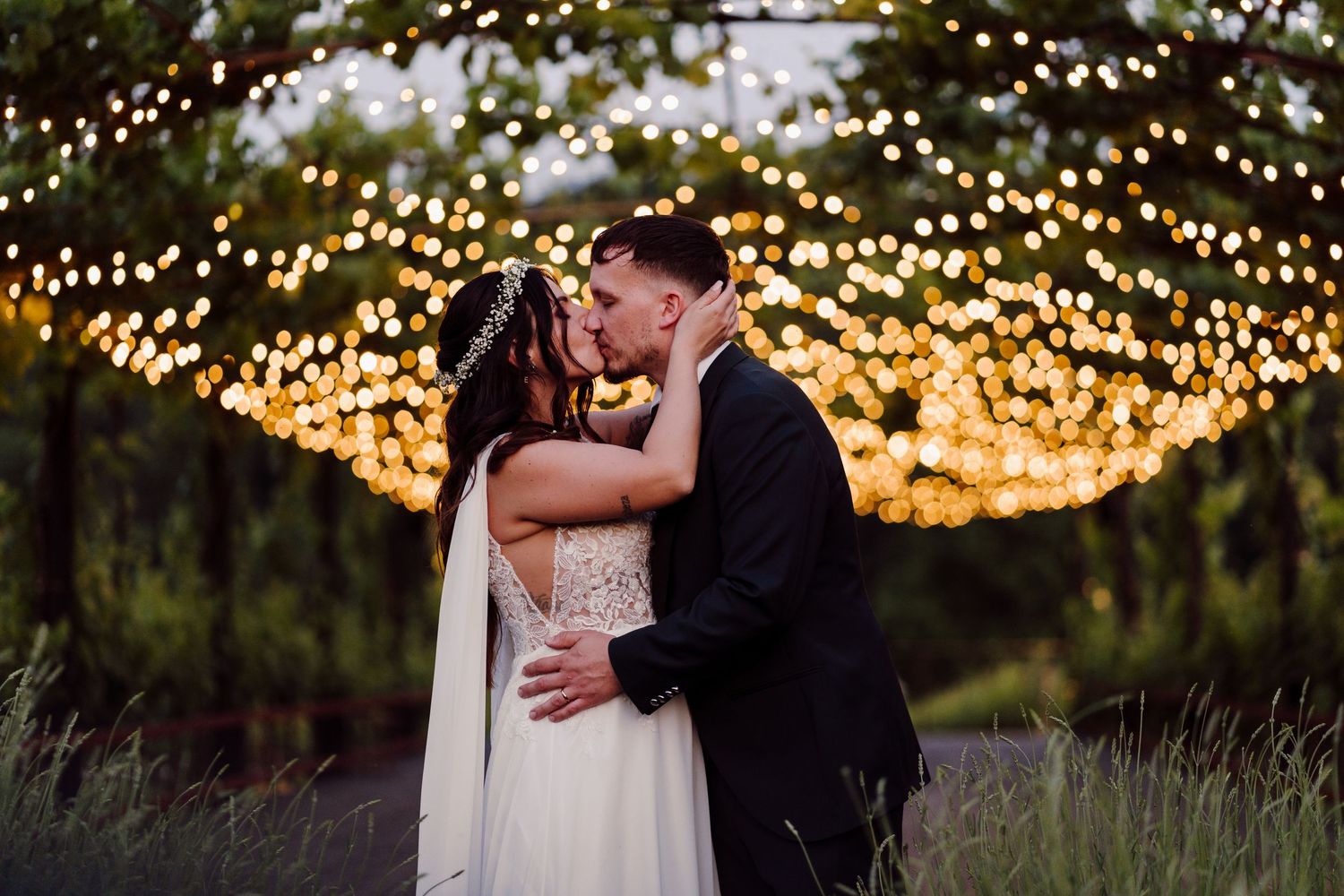 Bride and groom share a passionate kiss under a glowing canopy of fairy lights at twilight.