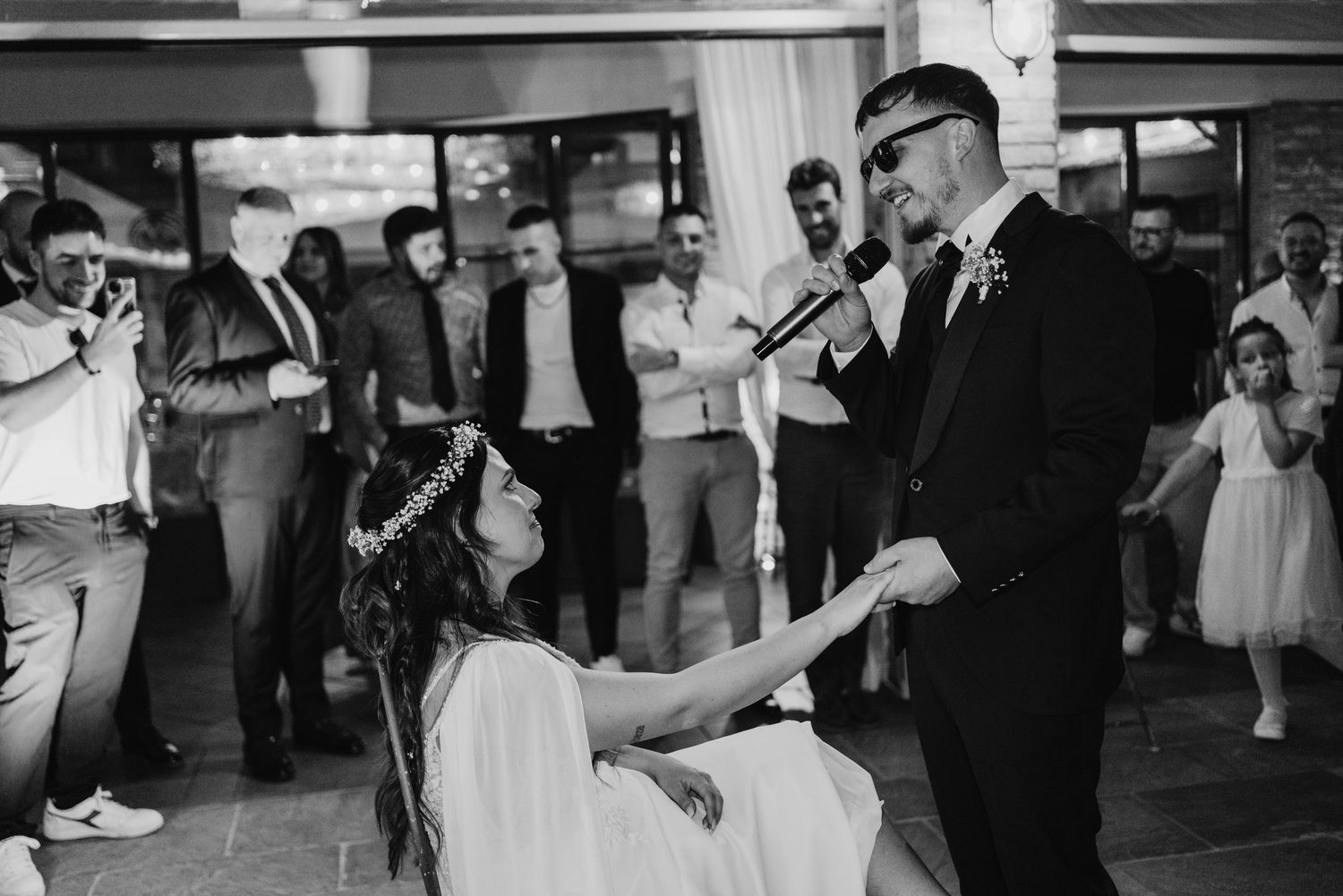 Black and white photo of groom serenading seated bride with microphone while wedding guests watch.