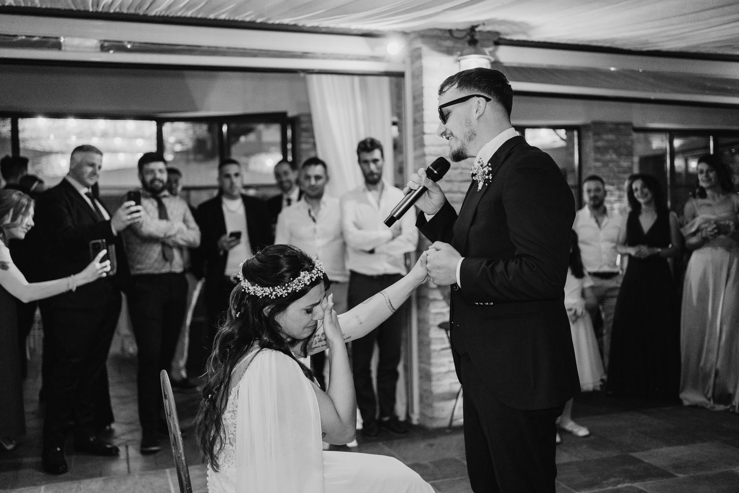 Black and white photo of groom singing into microphone while bride reaches up to touch his hand.