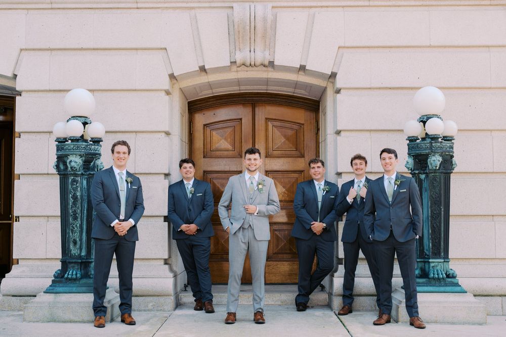 A wedding party of groomsmen in gray suits pose together on stone steps outside an elegant building.