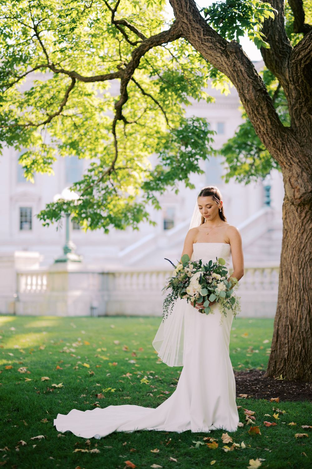 A bride in a white strapless wedding dress stands gracefully under green trees on a sunny day.