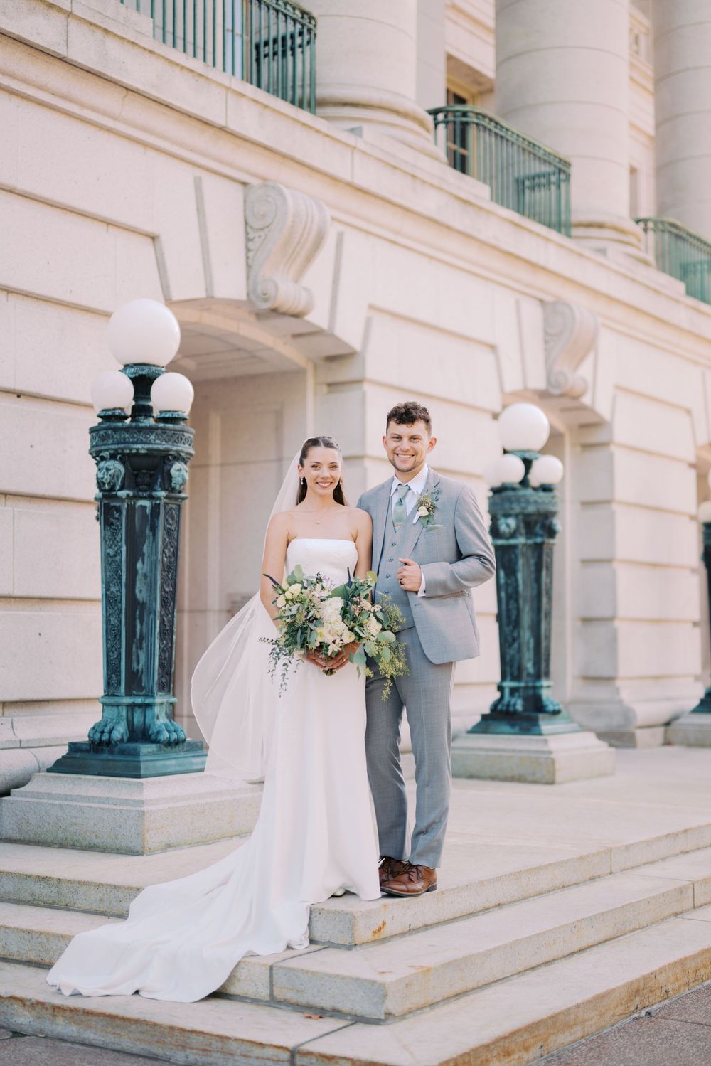 Elegant wedding couple poses on grand stone steps with ornate green lamp posts.