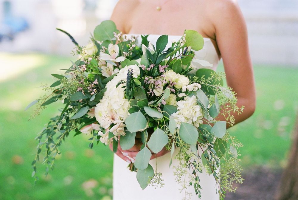 A delicate white and green wedding bouquet with eucalyptus leaves and roses against a soft outdoor background.