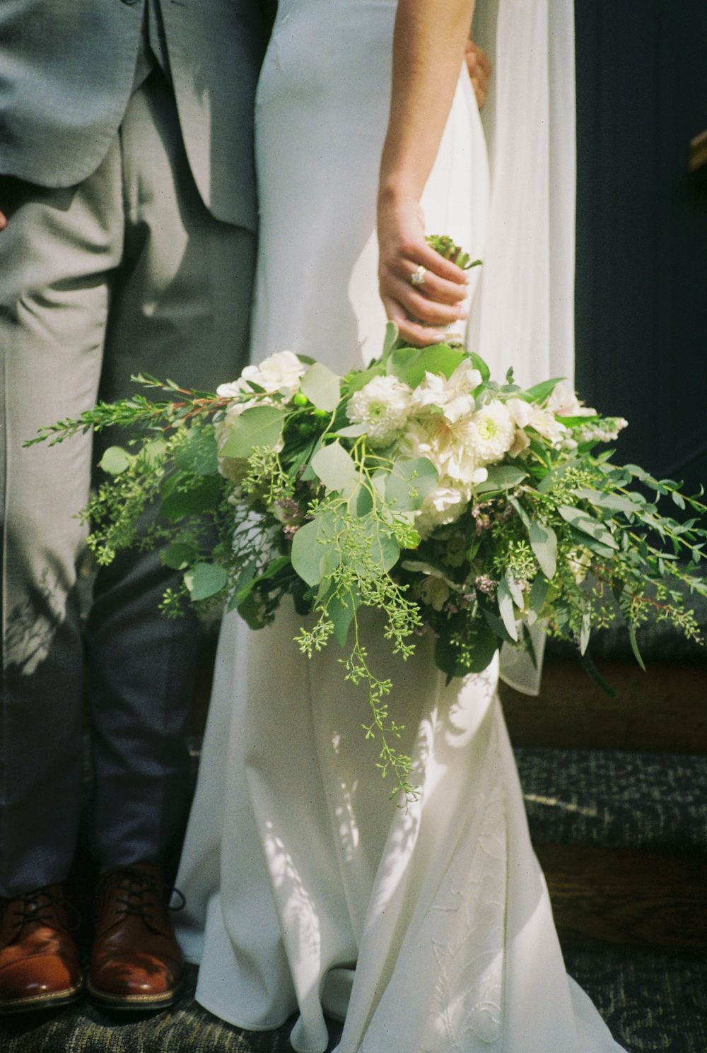 A wedding bouquet with white flowers and greenery held against a flowing white dress and gray suit.