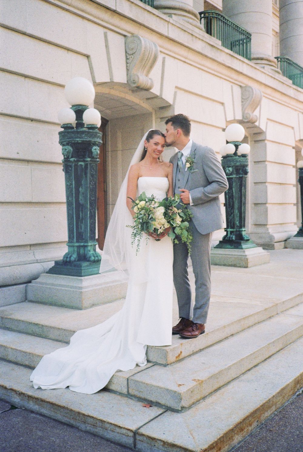 A couple shares a romantic moment on stone steps outside a grand building with ornate columns and architecture.