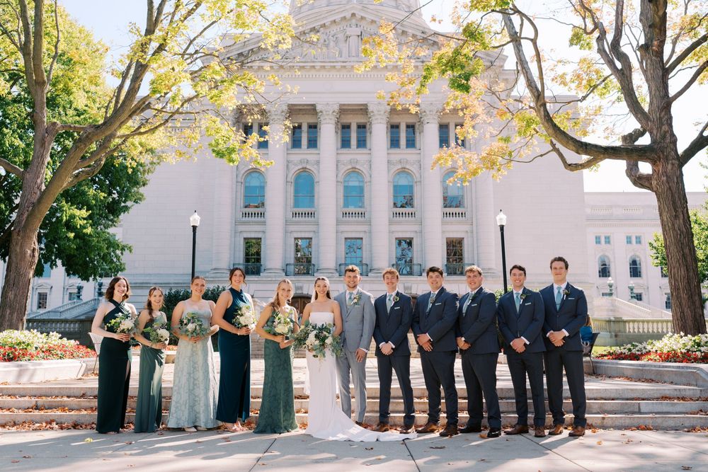 Wedding party in formal attire poses on grand steps of elegant white building with columns and blue accents.