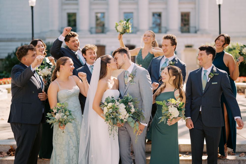 Wedding party in formal attire gathered outdoors for group photos with gray and sage green color scheme.