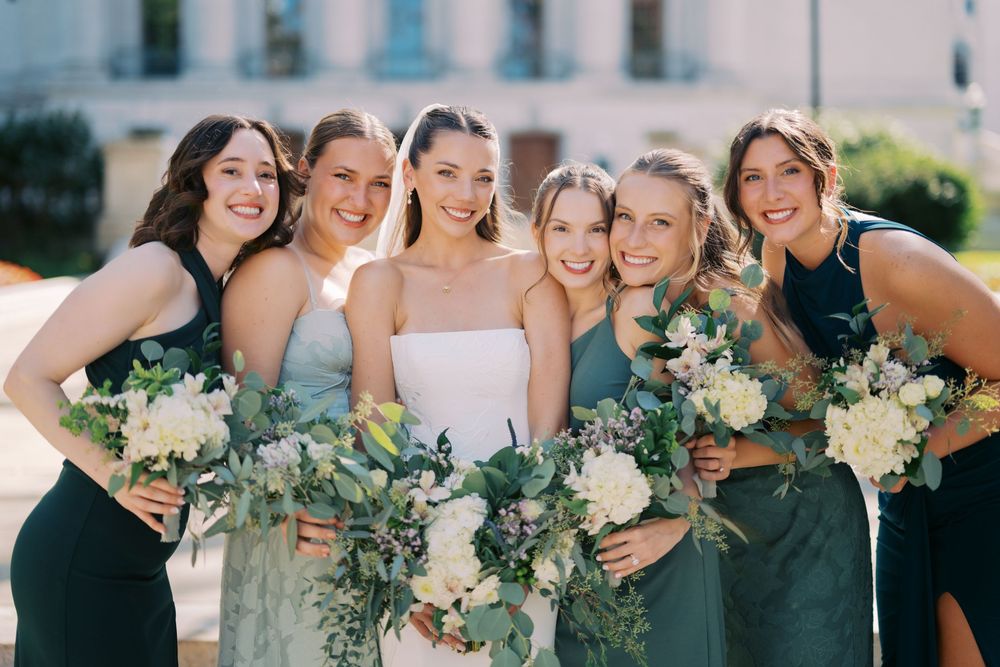 Bridal party holding white and green floral bouquets poses together outdoors on sunny day.
