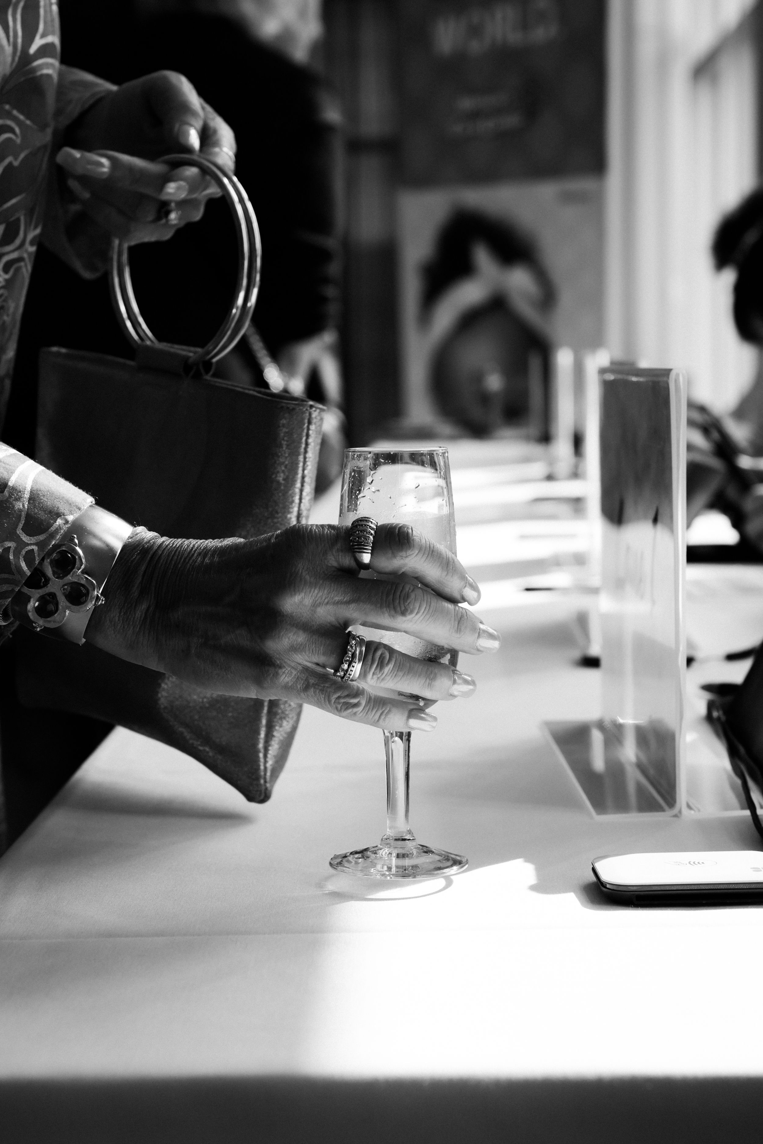 Close-up black and white image of a womans hands holding a champagne glass and handbag at an event.