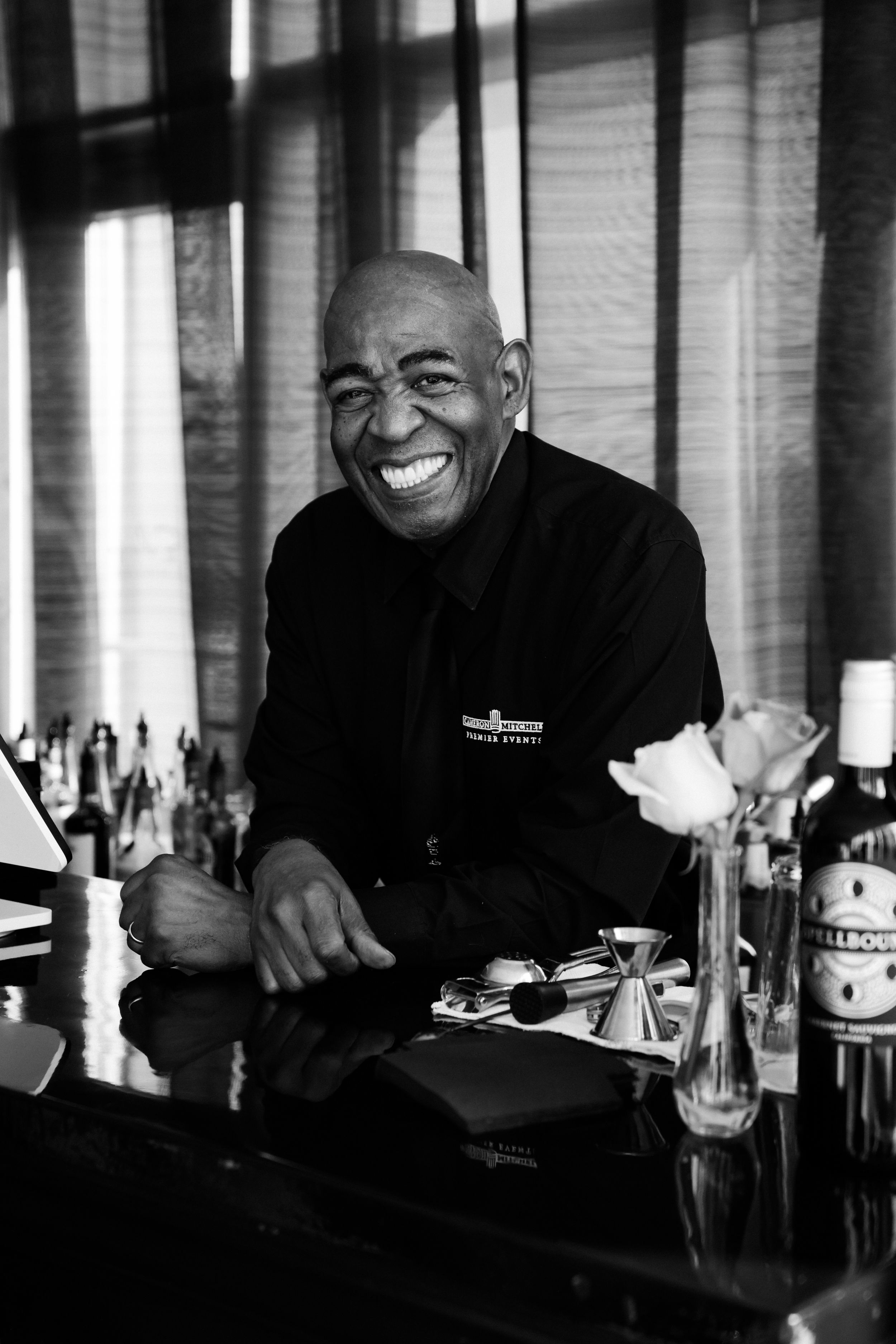 Joyful bartender smiling behind a bar counter in a black and white portrait at a formal event.